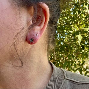 May include: A silver-toned star-shaped stud earring worn in the lower earlobe, accompanied by a small, thin, rectangular silver-toned stud earring positioned directly above it in a secondary piercing on the ear.