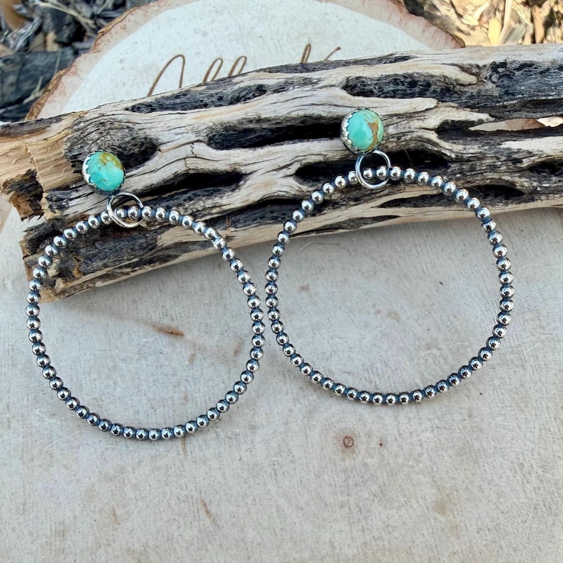 Puede incluir: Un par de pendientes de aro plateados con detalles de piedras de color turquesa. Los aros est&aacute;n hechos de peque&ntilde;as cuentas redondas. Los pendientes se exhiben sobre un trozo de madera.