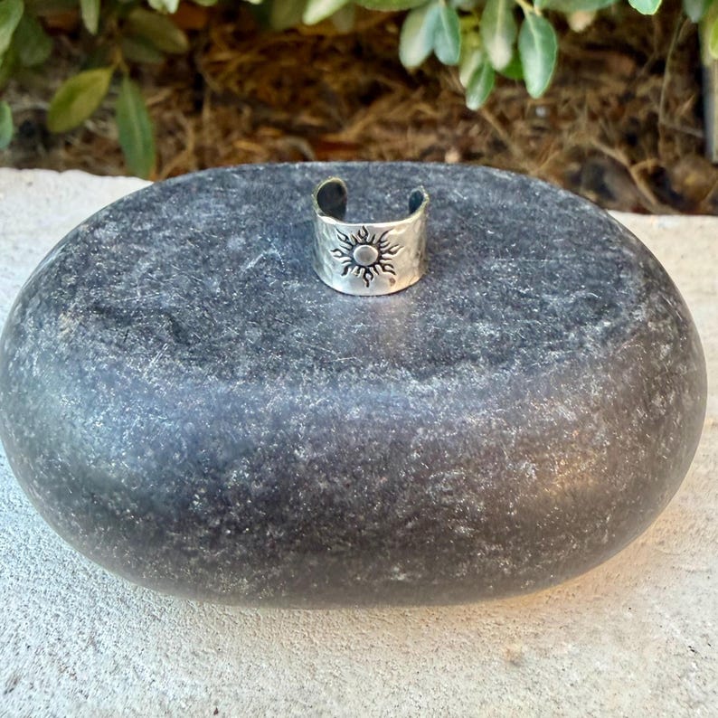 May include: A silver-toned metal ring featuring an open band design with a stamped sun motif. The surface of the ring has a textured, hammered finish, and the central sun emblem displays a circular core surrounded by radiating rays.