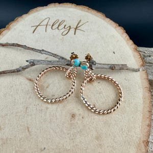 May include: A pair of gold-toned hoop earrings with a twisted rope design. Each earring features a small, round, turquoise stone. The earrings are displayed on a wooden surface with the handwritten text "Allyk".