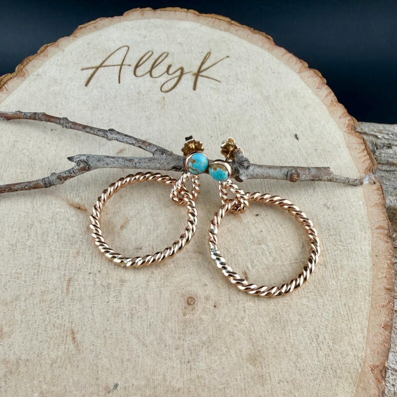 May include: A pair of gold-toned hoop earrings with a twisted rope design. Each earring features a small, round, turquoise stone. The earrings are displayed on a wooden surface with the handwritten text "Allyk".