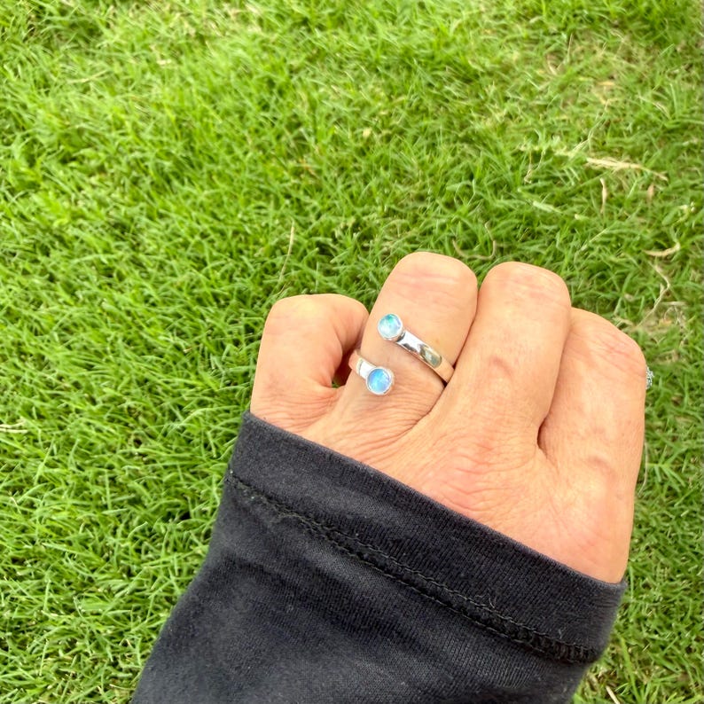 May include: A silver ring featuring two light blue gemstones. The ring is worn on a hand, with a black sleeve, against a backdrop of green grass. The gemstones are round and set in a minimalist design.