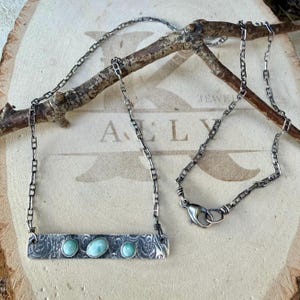 May include: A silver necklace featuring a rectangular bar pendant with an intricate etched pattern and three oval light blue stones. The pendant is attached to a delicate silver chain with a lobster claw clasp closure.