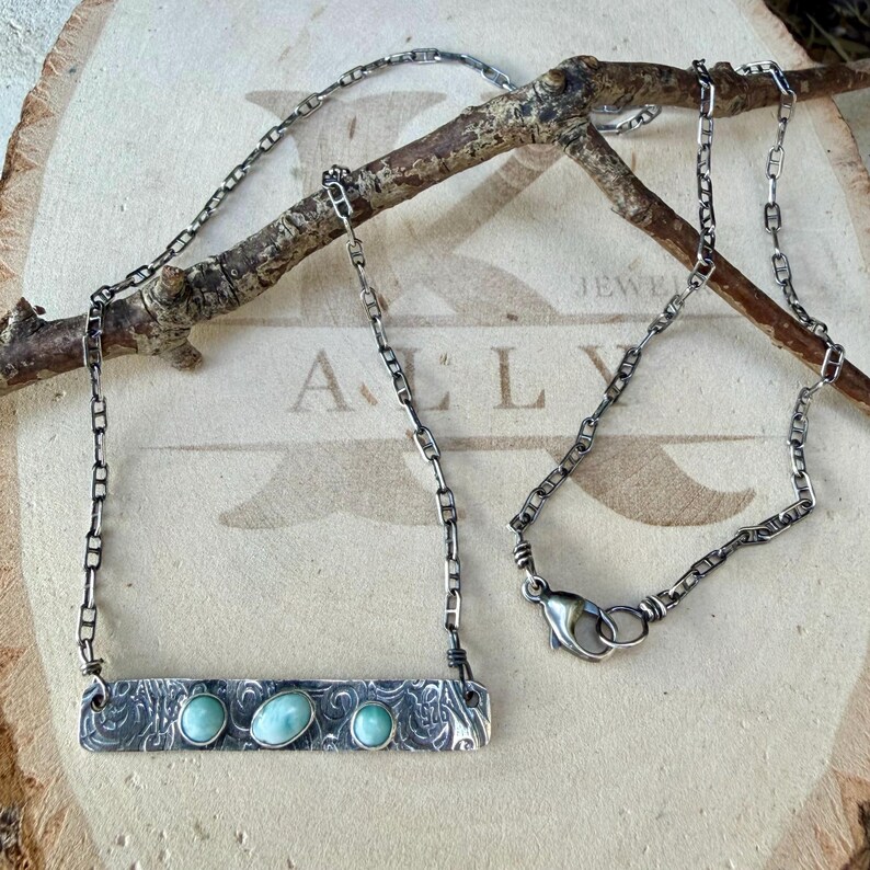 May include: A silver necklace featuring a rectangular bar pendant with an intricate etched pattern and three oval light blue stones. The pendant is attached to a delicate silver chain with a lobster claw clasp closure.