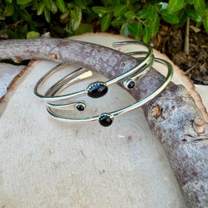 Op de afbeelding: Drie zilverkleurige armbanden met zwarte edelsteenaccenten. De open armbanden liggen op een licht houten oppervlak met een tak. De edelstenen zijn ovaal en rond.