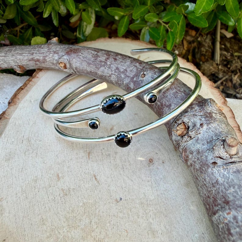 Op de afbeelding: Drie zilverkleurige armbanden met zwarte edelsteenaccenten. De open armbanden liggen op een licht houten oppervlak met een tak. De edelstenen zijn ovaal en rond.