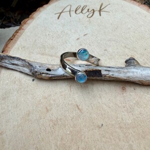 May include: A silver cuff bracelet featuring two iridescent blue-green stones. The open bracelet is curved, displayed on a piece of driftwood against a light wood background. The name "Ally K" is written in cursive.
