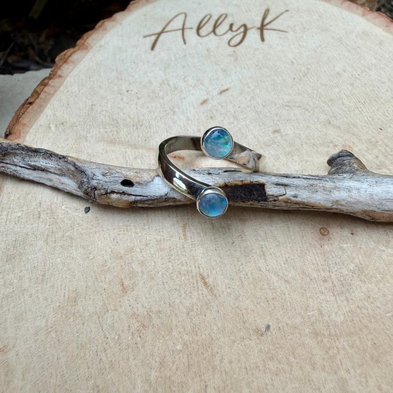 May include: A silver cuff bracelet featuring two iridescent blue-green stones. The open bracelet is curved, displayed on a piece of driftwood against a light wood background. The name "Ally K" is written in cursive.