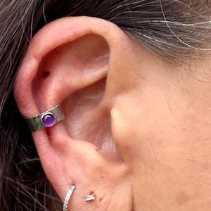 May include: A silver ear cuff with a textured band and a round, purple gemstone. The cuff is worn on the upper ear, with a small, sparkling earring visible below. The image is a close-up of the ear.