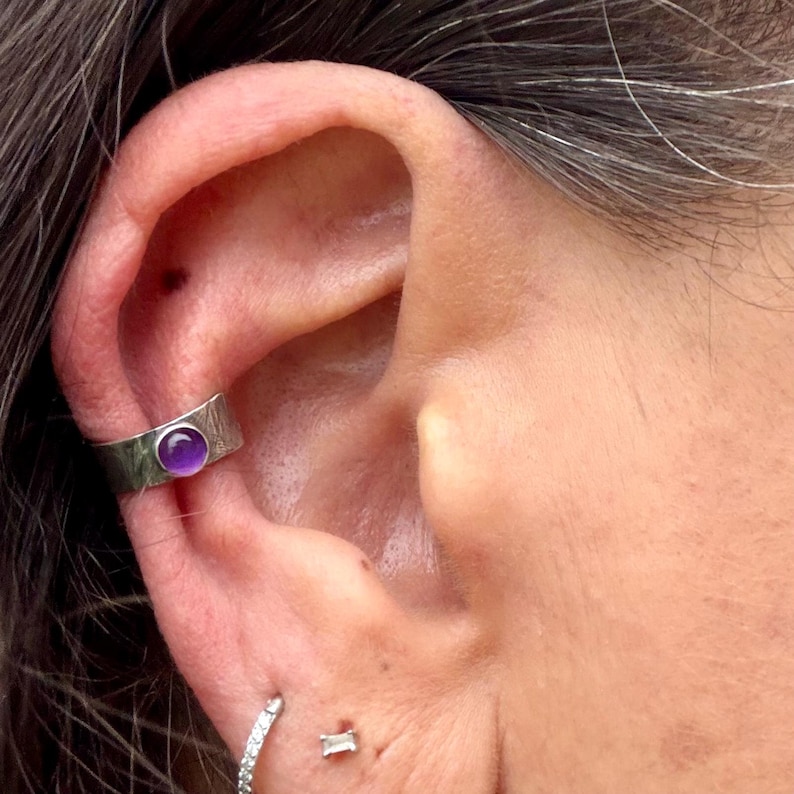 May include: A silver ear cuff with a textured band and a round, purple gemstone. The cuff is worn on the upper ear, with a small, sparkling earring visible below. The image is a close-up of the ear.