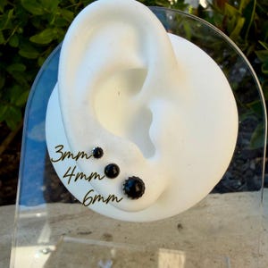 May include: Black stud earrings in 3mm, 4mm, and 6mm sizes are displayed on a white ear-shaped form. The sizes are labeled in black text. The earrings have a dark, possibly black, center stone.