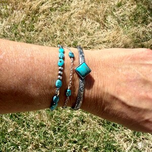 Puede incluir: Un conjunto de pulseras turquesas en una mu&ntilde;eca. Una pulsera presenta cuentas turquesas, otra tiene peque&ntilde;as piedras turquesas en una cadena, y la tercera es un brazalete de plata con una piedra turquesa cuadrada. Las pulseras se muestran sobre hierba verde.