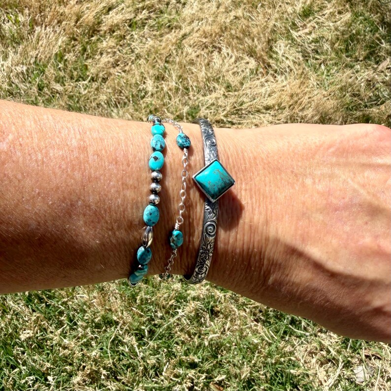 Puede incluir: Un conjunto de pulseras turquesas en una mu&ntilde;eca. Una pulsera presenta cuentas turquesas, otra tiene peque&ntilde;as piedras turquesas en una cadena, y la tercera es un brazalete de plata con una piedra turquesa cuadrada. Las pulseras se muestran sobre hierba verde.