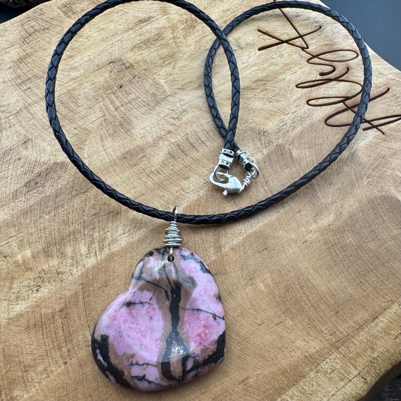 May include: A heart-shaped pendant necklace featuring a pink and black stone. The stone is wire-wrapped and suspended from a black braided leather cord with a silver-tone clasp. The necklace rests on a wooden surface.