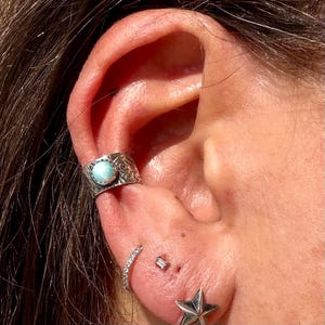 May include: A silver ear cuff featuring a round, light blue cabochon stone with an engraved floral pattern. Below it, a small silver hoop with clear stones, a tiny rectangular stud, and a silver star-shaped stud are worn in the earlobe.