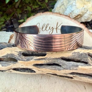 May include: A copper-colored cuff bracelet with a textured, engraved design. The bracelet has a dark interior and is displayed on a piece of driftwood. The design appears to be a topographical pattern.