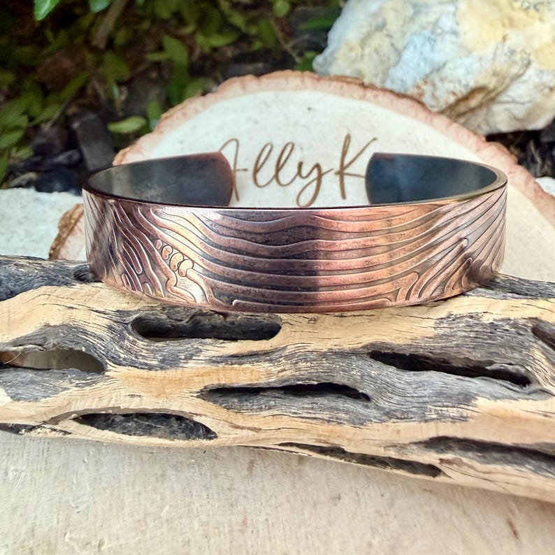 May include: A copper-colored cuff bracelet with a textured, engraved design. The bracelet has a dark interior and is displayed on a piece of driftwood. The design appears to be a topographical pattern.
