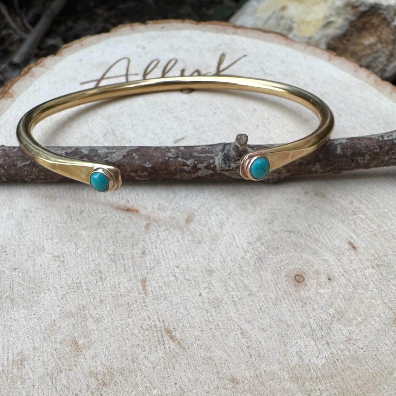 May include: A gold-colored open cuff bracelet with turquoise-colored stones at each end. The bracelet is resting on a wooden surface with the word "Allink" etched into it. The bracelet is approximately 2.5 inches in diameter.