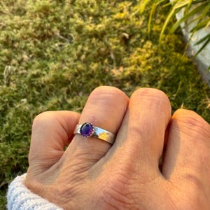 May include: A silver ring featuring a round, deep purple gemstone. The ring has a textured band with a floral design. The gemstone is set in a decorative, scalloped bezel. The ring is shown on a finger.