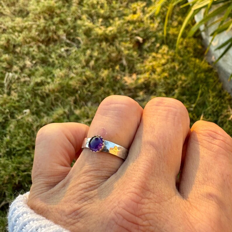 May include: A silver ring featuring a round, deep purple gemstone. The ring has a textured band with a floral design. The gemstone is set in a decorative, scalloped bezel. The ring is shown on a finger.