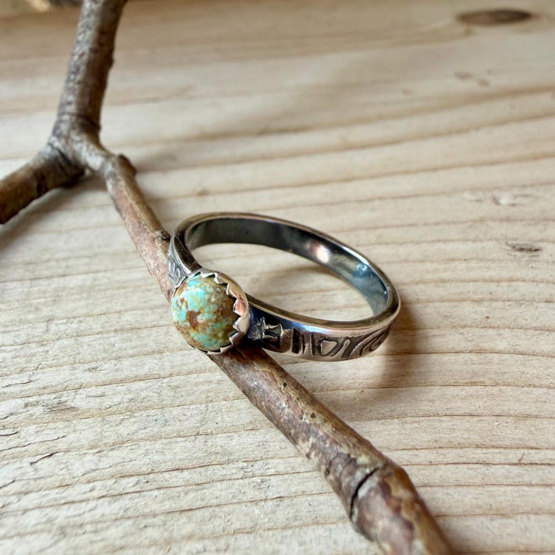 May include: A silver ring with a round, turquoise-colored stone. The ring band has a decorative pattern. The ring is resting on a small, brown twig against a wooden background.