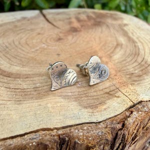 May include: A pair of silver heart-shaped stud earrings featuring an intricate, embossed scrollwork pattern across the surface. The metal has a darkened, oxidized finish within the recessed areas to highlight the detailed engraved design.
