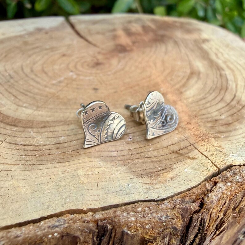 May include: A pair of silver heart-shaped stud earrings featuring an intricate, embossed scrollwork pattern across the surface. The metal has a darkened, oxidized finish within the recessed areas to highlight the detailed engraved design.