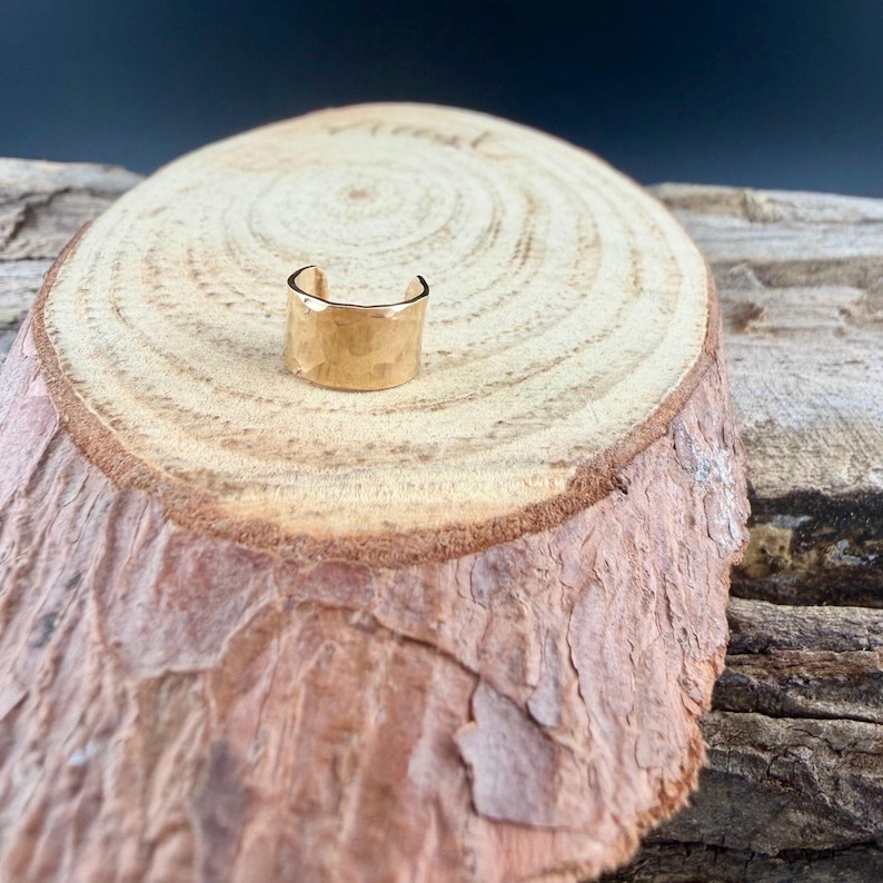 Puede incluir: Un brazalete de oreja dorado con textura martillada. El brazalete tiene un dise&ntilde;o sencillo y abierto y se muestra sobre una rodaja de madera con anillos de &aacute;rbol visibles.