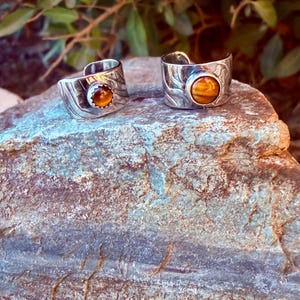 K&ouml;nnte beinhalten: Zwei silberfarbene Ringe mit offener Ringschiene, die jeweils mit einem runden, goldbraunen Tigerauge-Edelstein besetzt sind. Die Metallb&auml;nder weisen eine texturierte, wellenf&ouml;rmige Oberfl&auml;che auf. Die Ringe liegen nebeneinander.