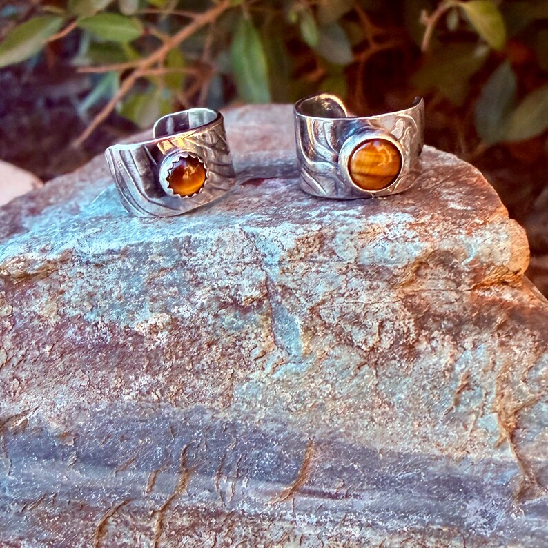 K&ouml;nnte beinhalten: Zwei silberfarbene Ringe mit offener Ringschiene, die jeweils mit einem runden, goldbraunen Tigerauge-Edelstein besetzt sind. Die Metallb&auml;nder weisen eine texturierte, wellenf&ouml;rmige Oberfl&auml;che auf. Die Ringe liegen nebeneinander.