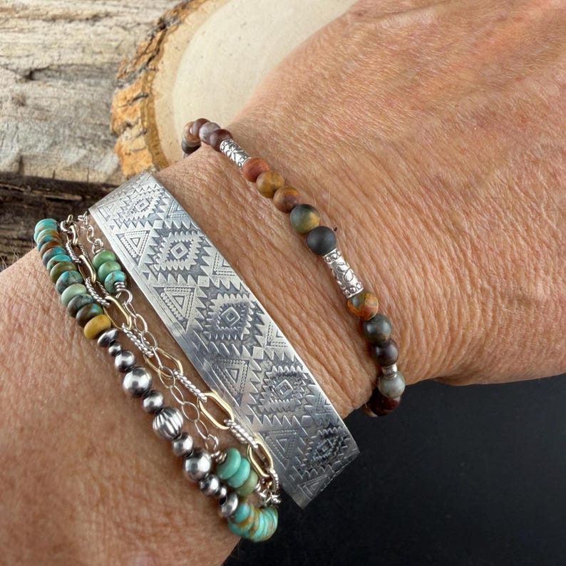 May include: A stack of three bracelets: one wide silver cuff with geometric engravings, one beaded bracelet with matte earth-toned stones and silver spacers, and one beaded bracelet featuring turquoise-colored discs and silver chain links.