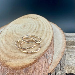 May include: Three gold-colored wavy rings are displayed on a wooden surface. The rings have a delicate, organic design, with a continuous wave pattern. The wood has a natural, light brown color with visible grain and a rough bark edge.