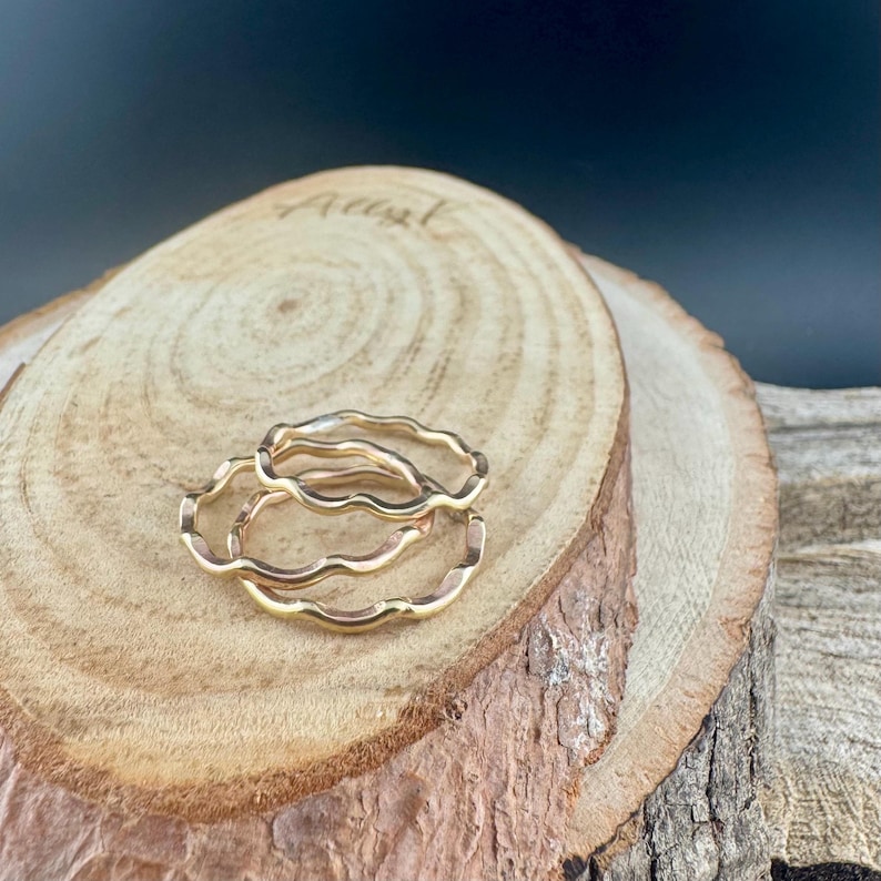 May include: Three gold-colored wavy rings are displayed on a wooden surface. The rings have a delicate, organic design, with a continuous wave pattern. The wood has a natural, light brown color with visible grain and a rough bark edge.