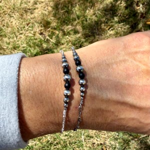 May include: Two beaded bracelets. One bracelet features a series of silver-colored beads interspersed with black beads. The other bracelet has a similar design. Both bracelets are on a silver chain.