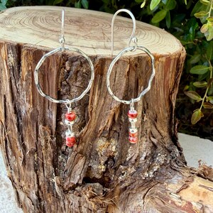 May include: A pair of silver dangle earrings featuring a large, hammered, circular hoop frame. Suspended from the center of each hoop is a vertical arrangement of three small, round, red and white marbled beads separated by tiny silver spacers.