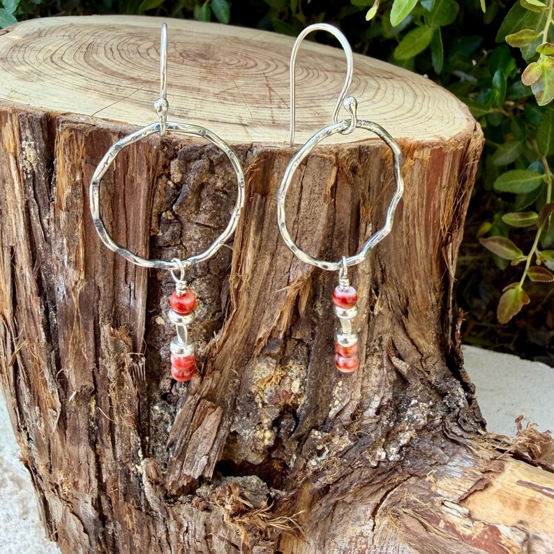 May include: A pair of silver dangle earrings featuring a large, hammered, circular hoop frame. Suspended from the center of each hoop is a vertical arrangement of three small, round, red and white marbled beads separated by tiny silver spacers.