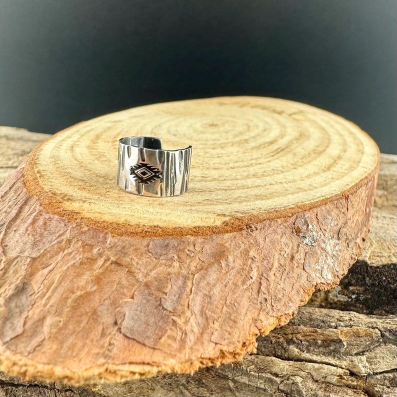 May include: A silver-colored ring with a geometric design. The ring has a textured, bark-like appearance and features a central diamond-shaped pattern with black accents. The ring is displayed on a wooden surface.