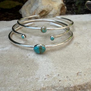 May include: Three silver-toned cuff bracelets with turquoise stones. The bracelets are simple, with a smooth, polished finish. The turquoise stones are oval-shaped and set in the center of each bracelet. The bracelets are stacked on top of each other.
