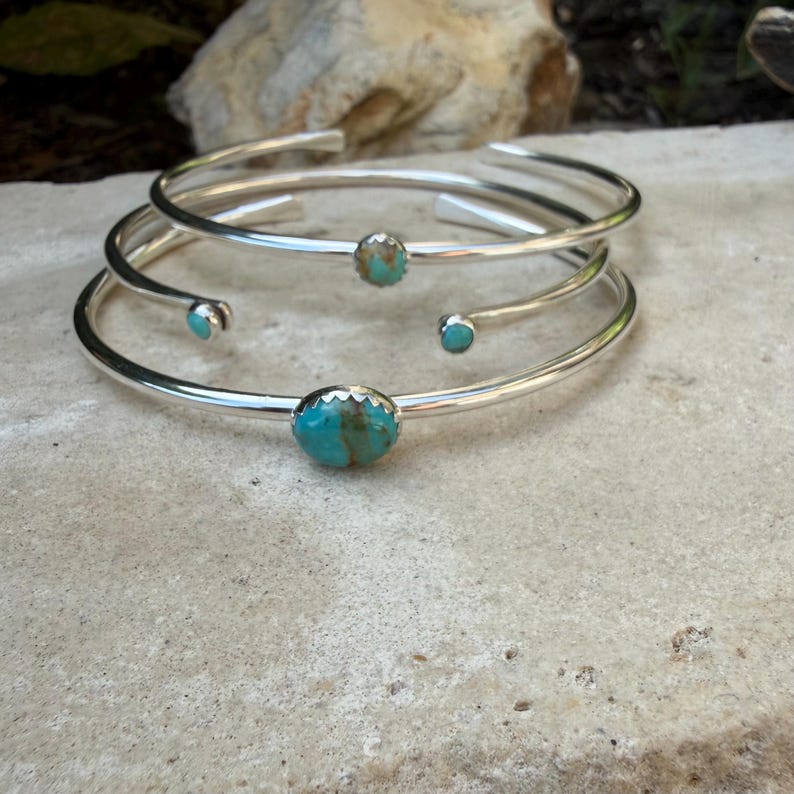 May include: Three silver-toned cuff bracelets with turquoise stones. The bracelets are simple, with a smooth, polished finish. The turquoise stones are oval-shaped and set in the center of each bracelet. The bracelets are stacked on top of each other.