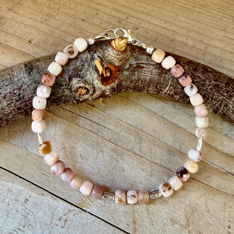 May include: A beaded bracelet featuring small, faceted, square-shaped beads in shades of pale pink, cream, and translucent brown. The bracelet is secured with a silver-toned metal lobster claw clasp and jump ring, creating a circular loop.