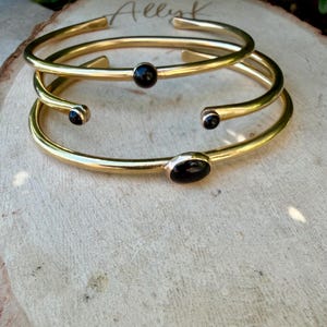 May include: Three gold-colored metal bracelets with black oval and round stones. The bracelets are stacked on a light brown wooden surface. The word "Allyk" is written in cursive on the wood.