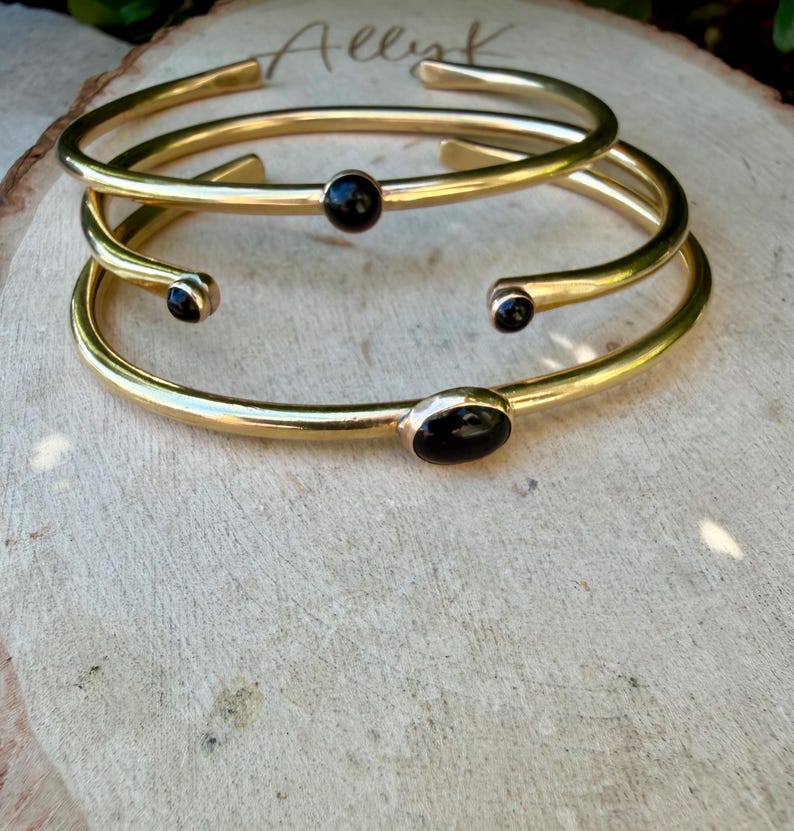 May include: Three gold-colored metal bracelets with black oval and round stones. The bracelets are stacked on a light brown wooden surface. The word "Allyk" is written in cursive on the wood.