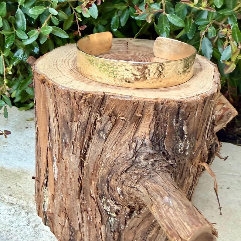 Puede incluir: Un brazalete tipo pu&ntilde;o de metal dorado con una superficie martillada y texturizada. La pulsera presenta un dise&ntilde;o abierto, lo que permite su ajuste. El metal tiene un acabado pulido y reflectante que capta la luz, mostrando las hendiduras irregulares creadas por el proceso de martillado en toda la banda exterior.