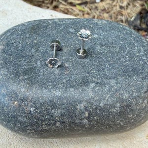 May include: A pair of silver-toned stud earrings featuring a small, detailed flower design at the top. Each earring has a straight post and a circular backing, showcasing a metallic finish with intricate petal detailing on the flower head.