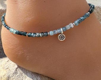 May include: Ankle bracelet featuring small, square-cut blue-green beads strung together with silver-colored accents. A small, silver-colored charm with a floral design dangles from the bracelet. The jewelry is designed to be worn around the ankle.