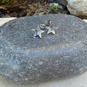 May include: A pair of silver-toned star-shaped stud earrings with a polished metallic finish. Each earring features a five-pointed star design with a secure post backing, showcasing a smooth, reflective surface that catches the light.