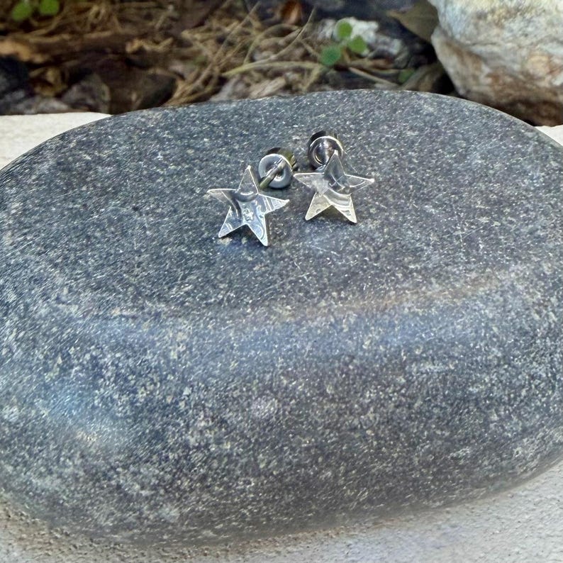 May include: A pair of silver-toned star-shaped stud earrings with a polished metallic finish. Each earring features a five-pointed star design with a secure post backing, showcasing a smooth, reflective surface that catches the light.