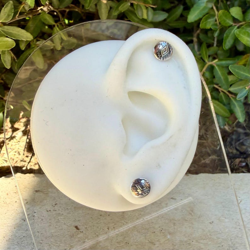 May include: A pair of small, round, silver-toned stud earrings featuring a centered diamond-shaped engraving with radiating lines. The earrings are displayed on a white, ear-shaped mannequin stand made of smooth, matte material.