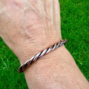 May include: A copper-colored twisted metal bracelet. The bracelet is worn on a wrist, with the twisted design creating a textured appearance. The bracelet's color is a mix of copper and darker tones.