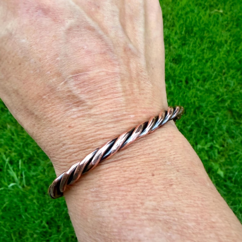 May include: A copper-colored twisted metal bracelet. The bracelet is worn on a wrist, with the twisted design creating a textured appearance. The bracelet's color is a mix of copper and darker tones.